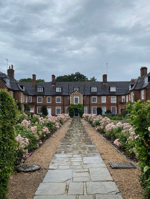 A symmetrical brick building with multiple windows and a central entrance surrounded by lush greenery, viewed from a paved stone pathway lined with blooming pink and white roses on both sides. The gar