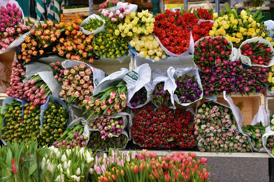 A vibrant display of freshly cut flowers arranged in bouquets and bundles at a flower market, featuring tulips in shades of pink, white, and red at the front, with larger bouquets of mixed flowers inc
