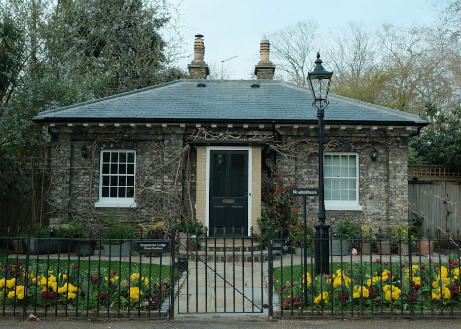 A quaint brick cottage with a dark blue front door and two white-framed windows, surrounded by a black metal fence and a garden filled with vibrant yellow and red flowers, with a vintage-style lamp po