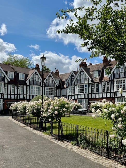 A row of traditional terraced houses with black and white timber-framed facades and steep gabled roofs, set behind a garden enclosed by a black metal fence. In the foreground, there are lush white flo