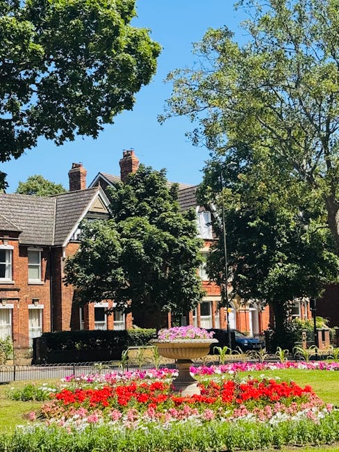 A vibrant flower bed with densely planted red, pink, and white blooms in front of a historic brick building with pitched roofs and chimneys, surrounded by lush green trees under a bright blue sky. The