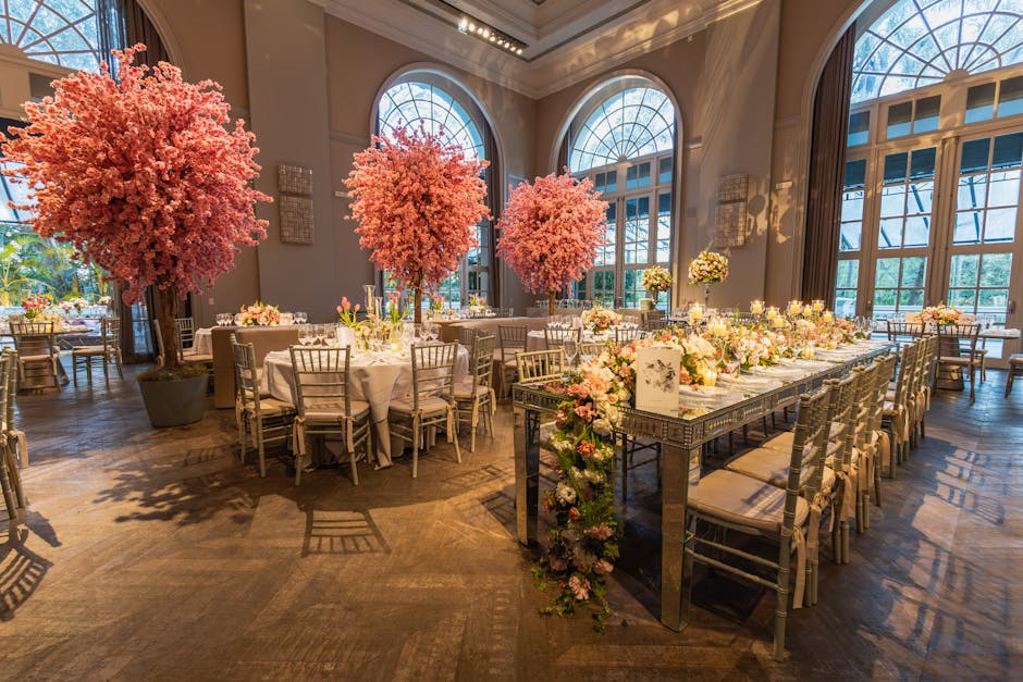 An elegantly decorated event space featuring tall, lush pink cherry blossom trees in large pots, positioned behind round tables draped with white tablecloths. The tables are adorned with delicate flor