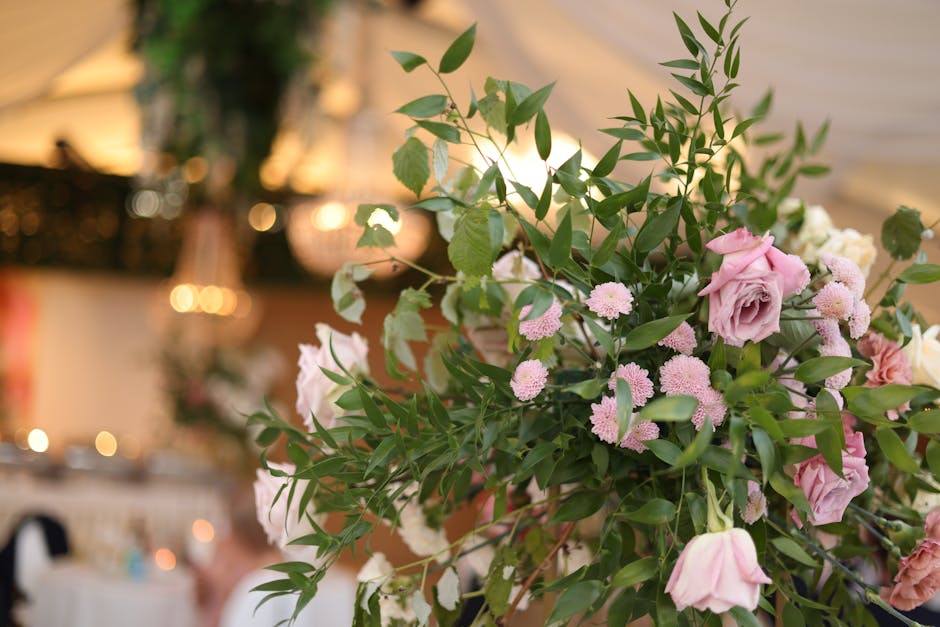 A large floral arrangement featuring a mix of soft pink roses, small pink and white chrysanthemum-like flowers, and lush green foliage arranged in a natural, slightly cascading style. The bouquet is s